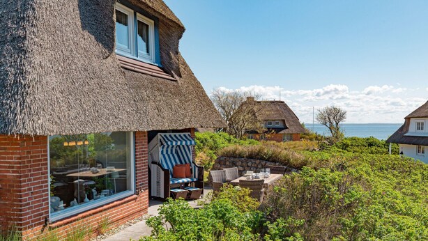 Ein Haus mit Strohdach, einer Glastür und einer Terrasse mit Gartenmöbeln.