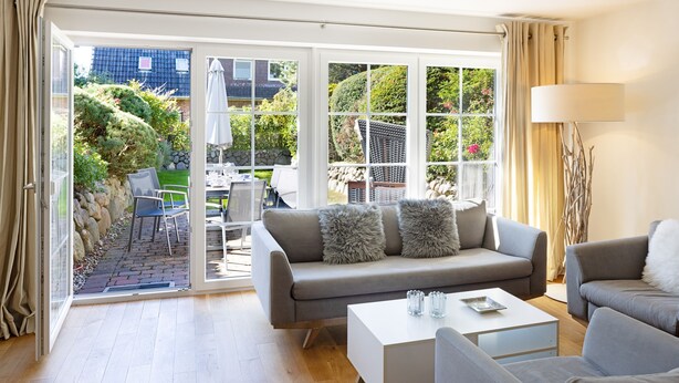 Una sala de estar moderna con un sofá gris, una mesa de centro blanca y un sillón gris. Se ve una ventana grande con vistas a un jardín y un edificio al fondo.