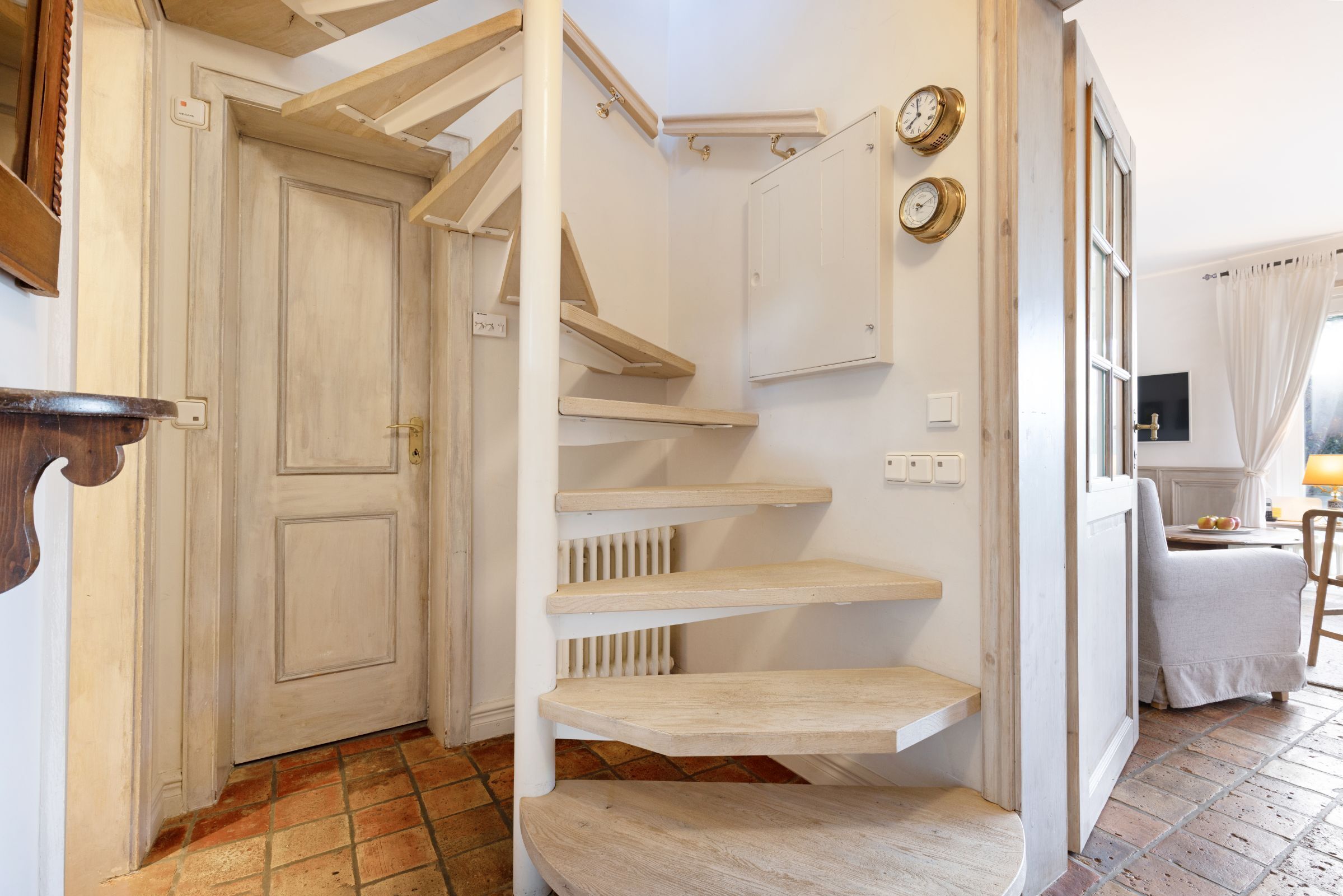 Una escalera de madera con peldaños que lleva a una plataforma, una puerta blanca y una pequeña mesa con una lámpara al fondo.