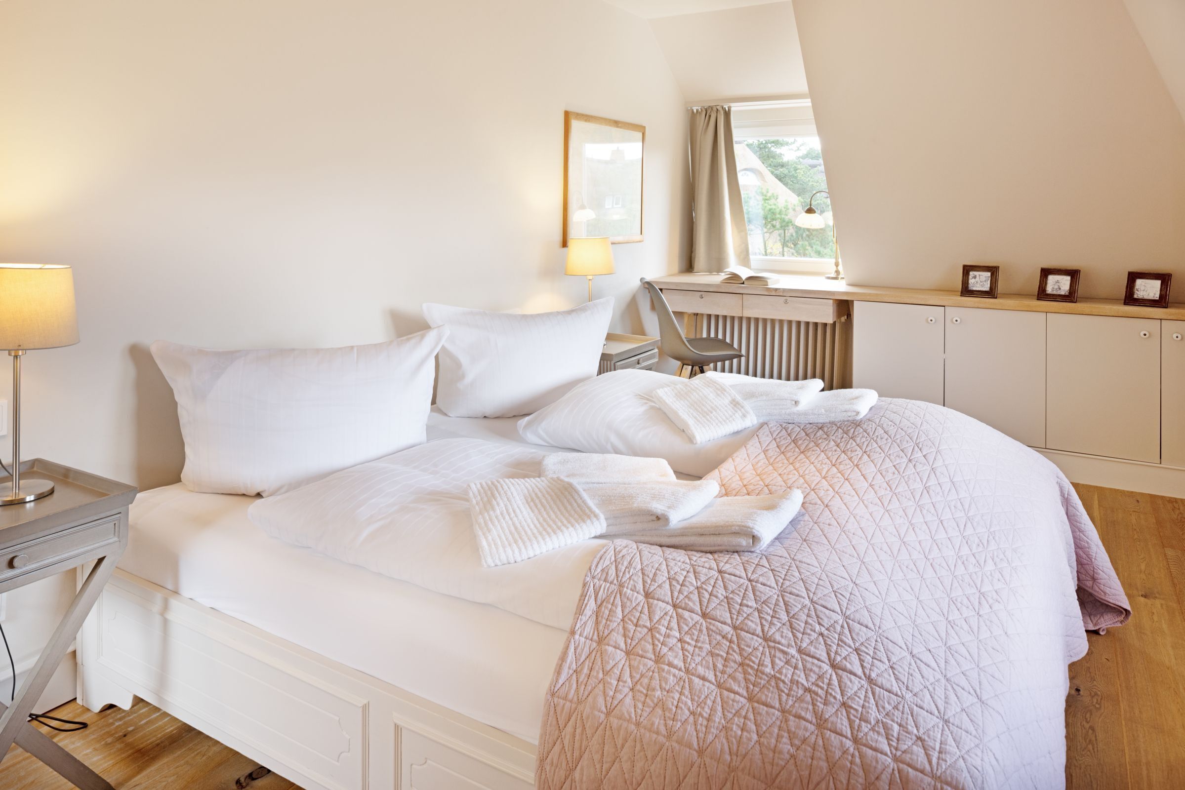 Una cama bien hecha con almohadas y una colcha, una lámpara de noche, un escritorio con silla y una ventana con cortinas.