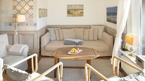 Un salón acogedor con un sofá beige, dos sillas de madera, una mesa redonda con fruta, una lámpara y un cuadro en la pared.