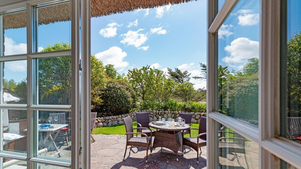Eine offene Terrasse mit rundem Tisch und Stühlen, Steinboden und Blick auf einen Garten mit Bäumen und einer Steinmauer.