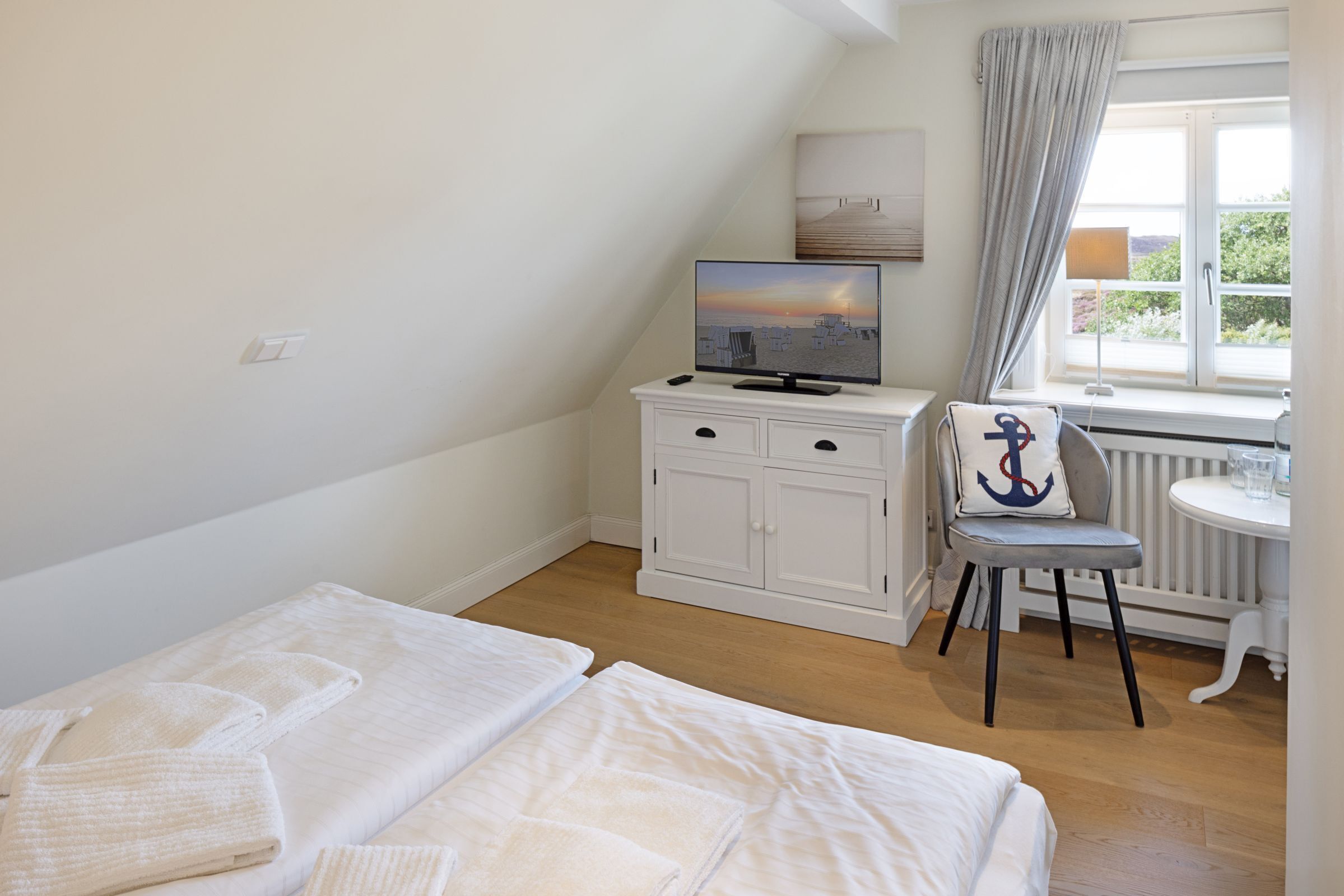 Een slaapkamer met twee bedden, een televisie op een witte kast, een stoel met een kussen, en een raam met gordijnen.