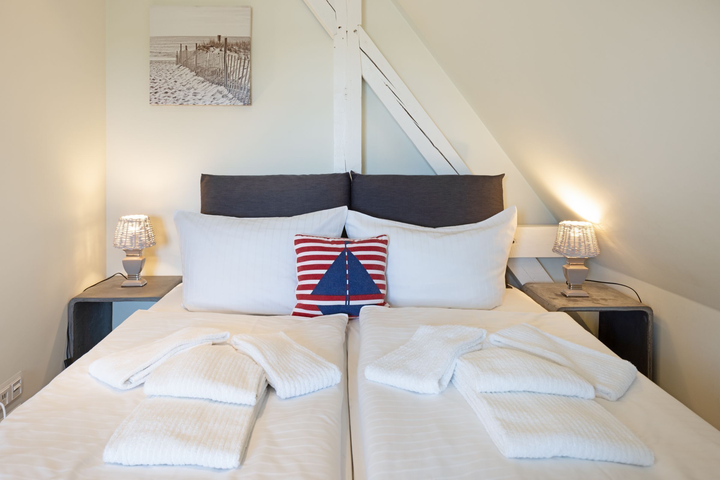 Een tweepersoonsbed met witte lakens, twee bedlampjes, een decoratief kussen met streepdessin en een aan de muur hangend schilderij.