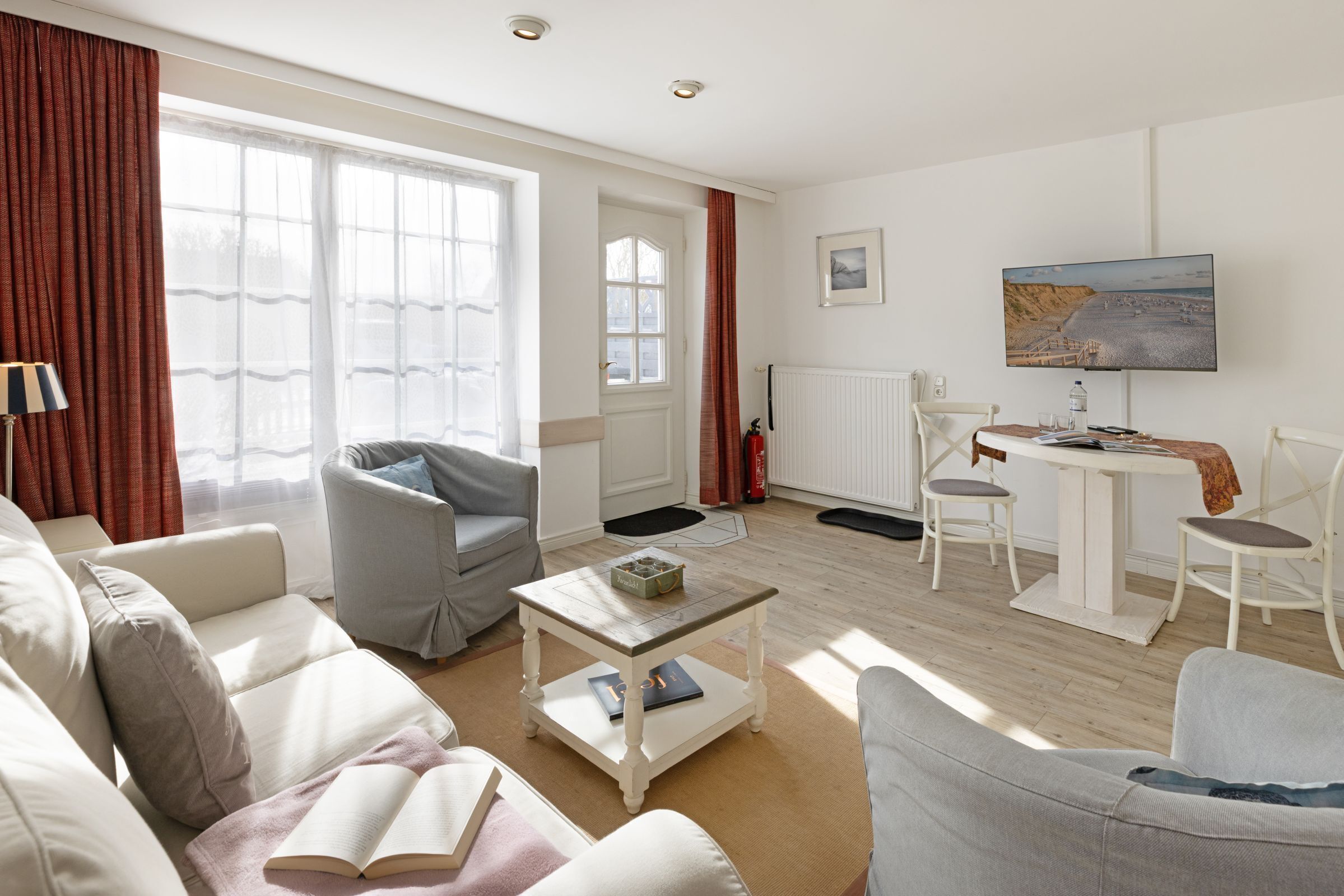 A spacious living area with a sofa, armchairs, a coffee table, and a dining table. There is a large window with curtains, a radiator, and a painting on the wall.