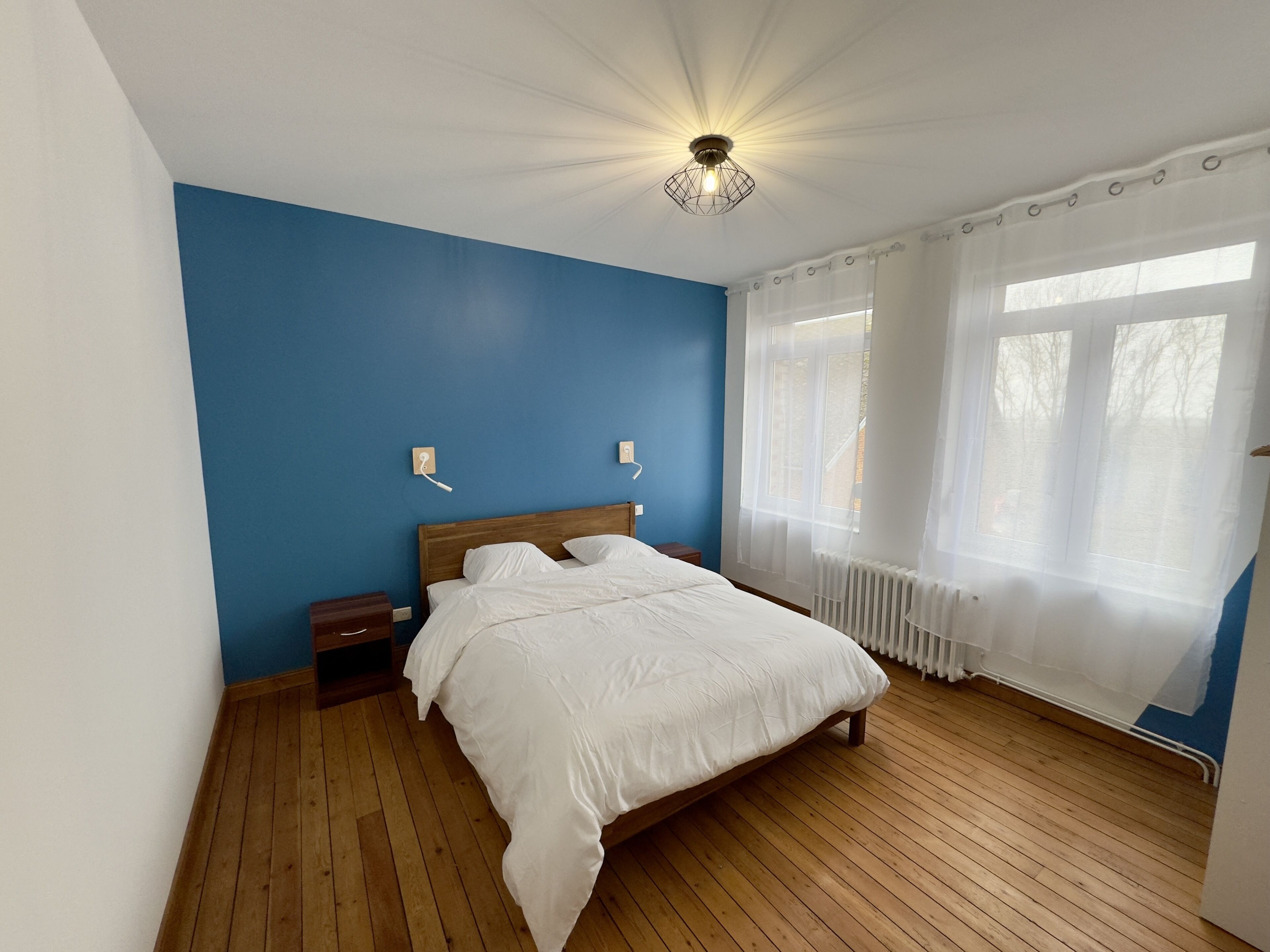 Quarto com piso de madeira, cama com roupa de cama branca, mesa de cabeceira, luminária na parede e janela com cortinas leves.