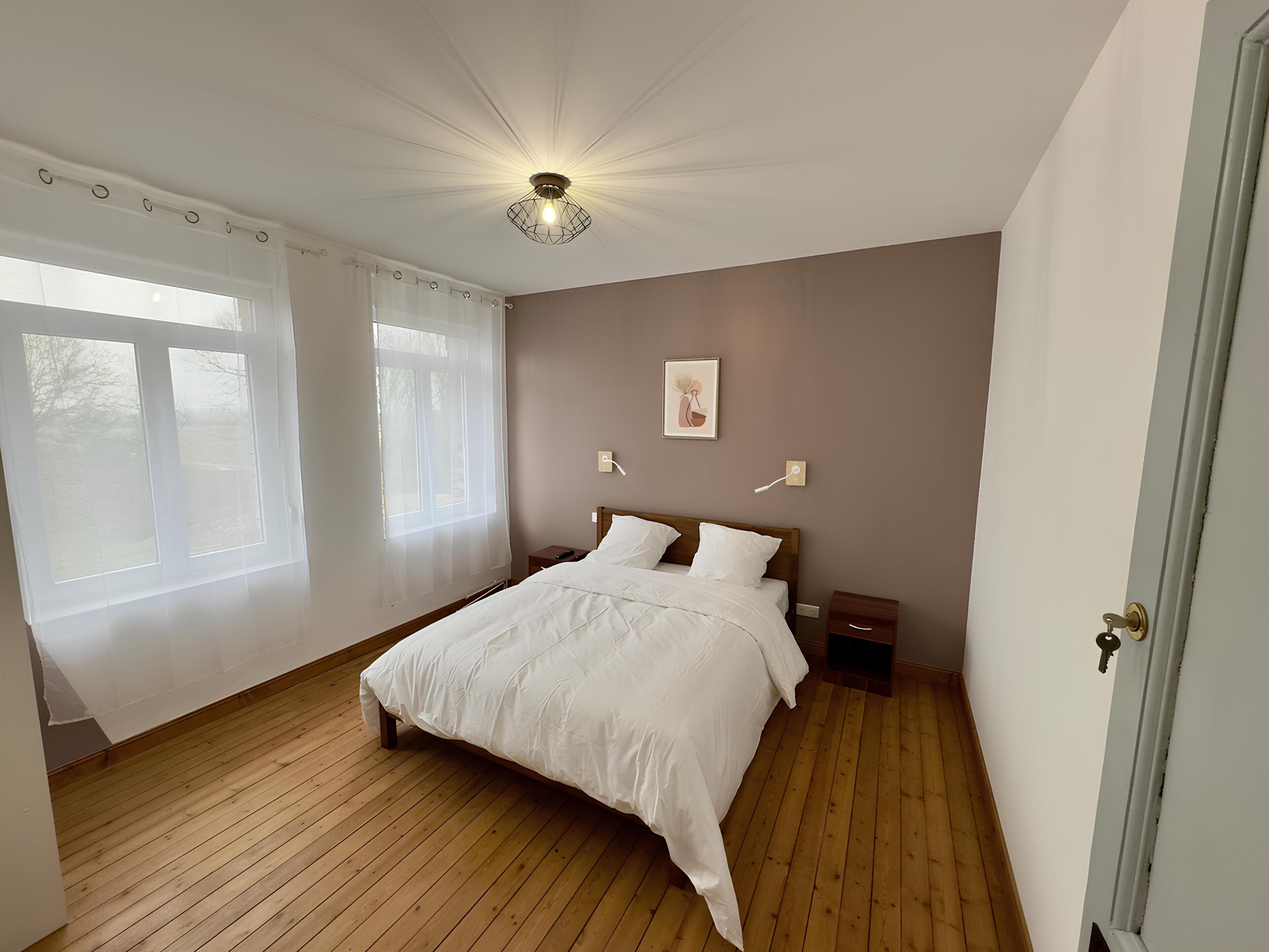 Ein Schlafzimmer mit Holzboden, einem Bett mit weißer Bettwäsche, einem Nachttisch, einer Wandleuchte und einem Fenster mit durchscheinenden Vorhängen.