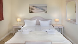 Una cama bien hecha con sábanas y almohadas blancas, dos mesitas de noche con lámparas, un espejo grande en la pared y un cuadro enmarcado sobre la cama.