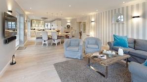 Un salon moderne avec un canapé gris, deux fauteuils, une table basse et un coin repas agrémenté d’un lustre.
