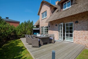 Une maison avec une façade en briques, un toit de chaume et une terrasse en bois aménagée avec du mobilier d’extérieur.