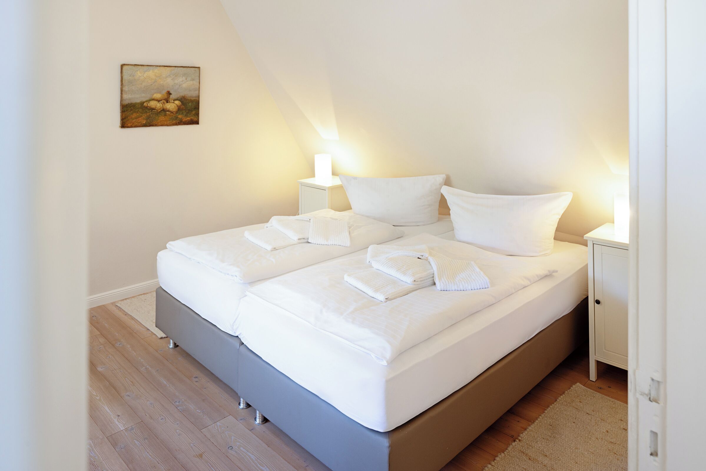 A neatly made bed with white linens and pillows, a bedside table with a lamp, and a painting on the wall.