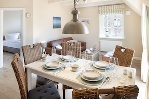 A dining area with a table set for six, surrounded by wicker chairs, a hanging light fixture, and a view of the living room in the background. - Herzlich Willkommen im Appartement Ankerplatz (List)