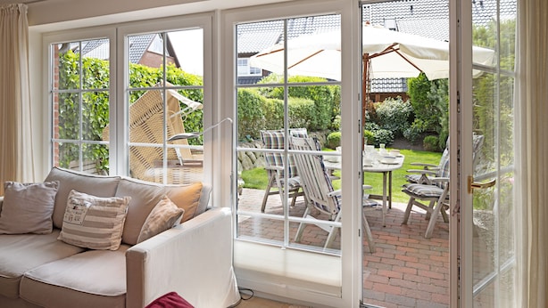 A room with a large sliding glass door leading to an outdoor patio area. Inside, there is a white sofa with striped pillows and a red chair.