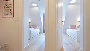 A bedroom with a white bed, a bedside table, and a nightstand. There is a framed picture on the wall and a plant on the floor.