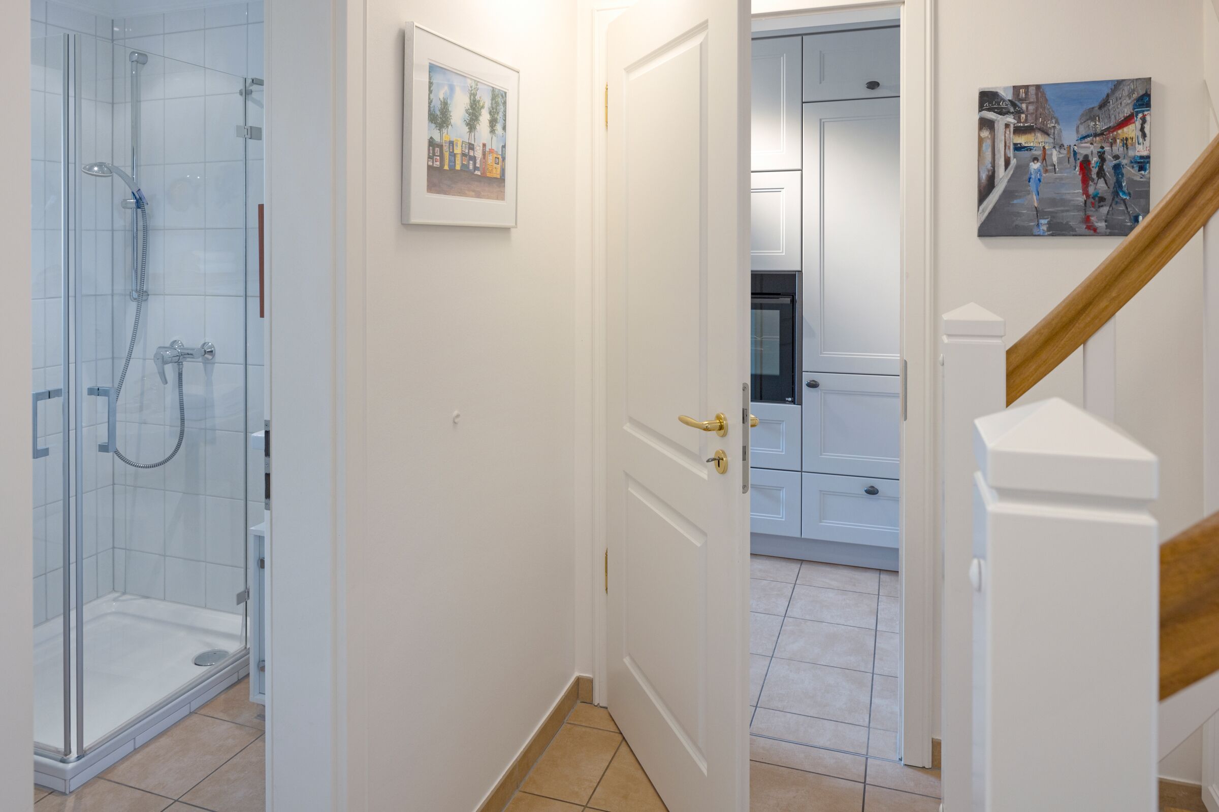 A modern bathroom with a glass shower enclosure, a white door with a gold handle, and a hallway leading to a kitchen area.
