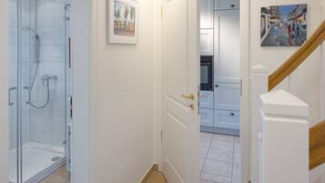 A modern bathroom with a glass shower enclosure, a white door with a gold handle, and a hallway leading to a kitchen area.
