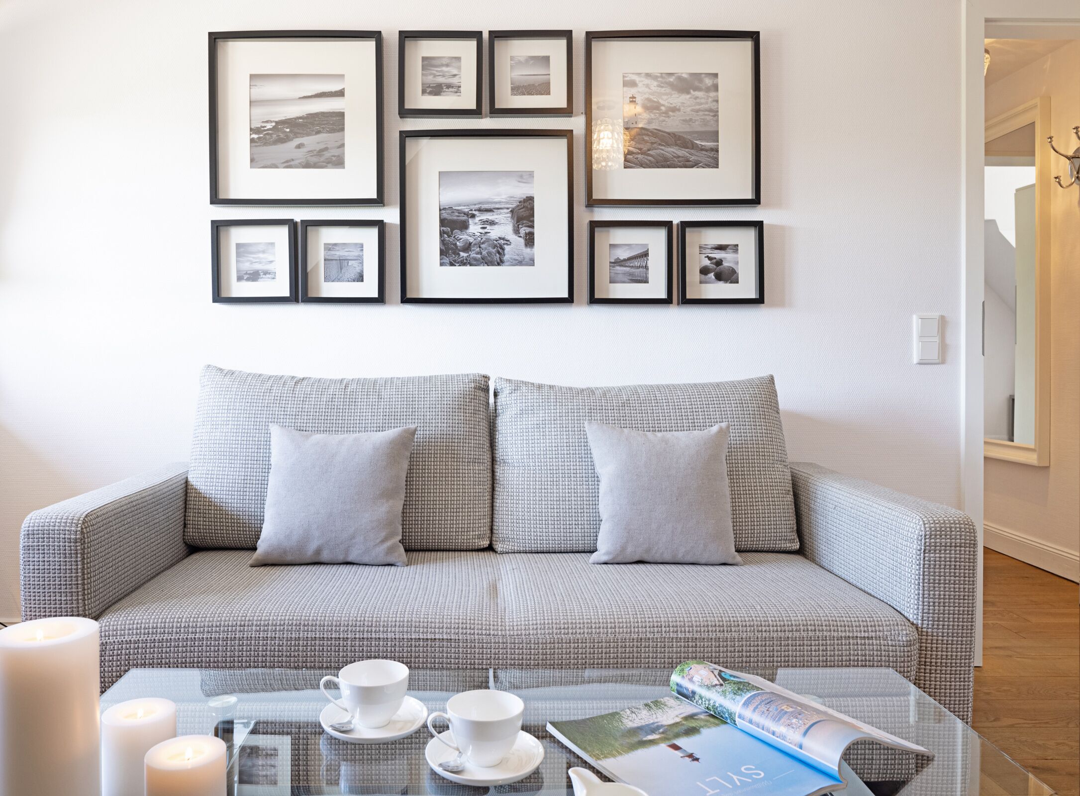 Una sala de estar con un sofá gris estampado, una mesa de centro de vidrio con tazas y una revista, y una pared decorada con fotografías en blanco y negro enmarcadas.