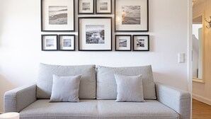 Un salon avec un canapé gris à motifs, une table basse en verre sur laquelle reposent des tasses et un magazine, et un mur décoré de photographies en noir et blanc encadrées.