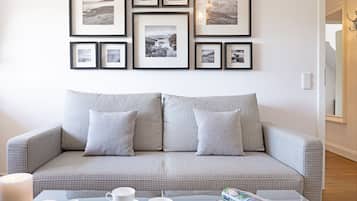 Una sala de estar con un sofá gris estampado, una mesa de centro de vidrio con tazas y una revista, y una pared decorada con fotografías en blanco y negro enmarcadas.