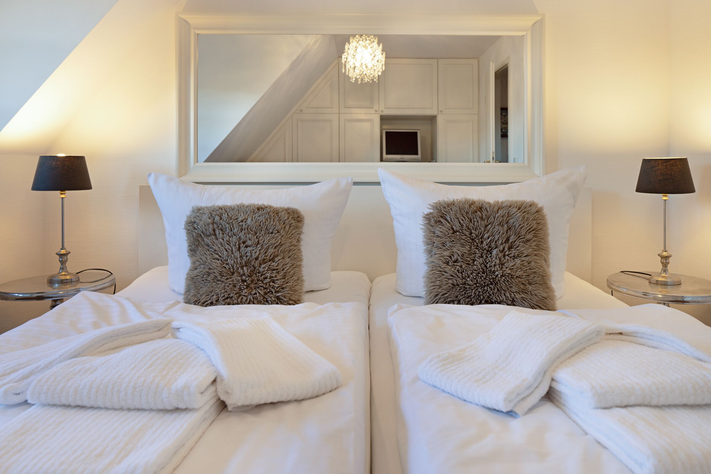 Hotelkamer met twee bedden, witte beddengoed, zachte kussens en een grote spiegel die een trap en een kroonluchter weerspiegelt.