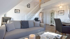 Ein modernes Wohnzimmer mit einem grau gemusterten Sofa, einem Glastisch mit Kerzen und einer Treppe mit weißen Geländern, die zu einer oberen Ebene führt.