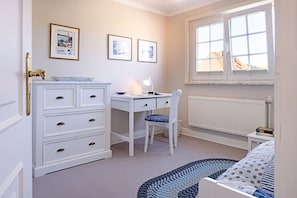 A bedroom with a white dresser, a desk with a chair, a window with a view, and a bed with a blue and white patterned bedspread.