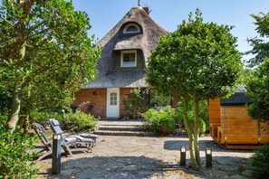 A thatched-roof cottage with a brick entrance, a small porch with a bench, and a wooden structure to the right.