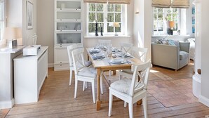 Uma sala de jantar com uma mesa de madeira posta para quatro pessoas, rodeada por cadeiras brancas. Há uma janela com vista para árvores e um armário branco com portas de vidro.