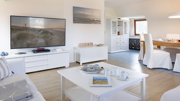 Una sala de estar moderna y bien iluminada, con un televisor de pantalla plana, un comedor con mesa de madera y una cocina visible al fondo.