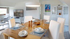 Un comedor con una mesa de madera dispuesta para seis personas, sillas blancas, un televisor y un aparador con puertas de cristal.