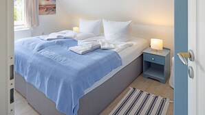 Un dormitorio con cama, almohadas, una mesita de noche con lámpara, una ventana con cortinas y un cuadro colgado en la pared.