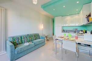 Una sala de estar moderna con un sofá azul claro, una mesa de comedor con sillas, y una cocina con armarios blancos y techo color verde azulado.