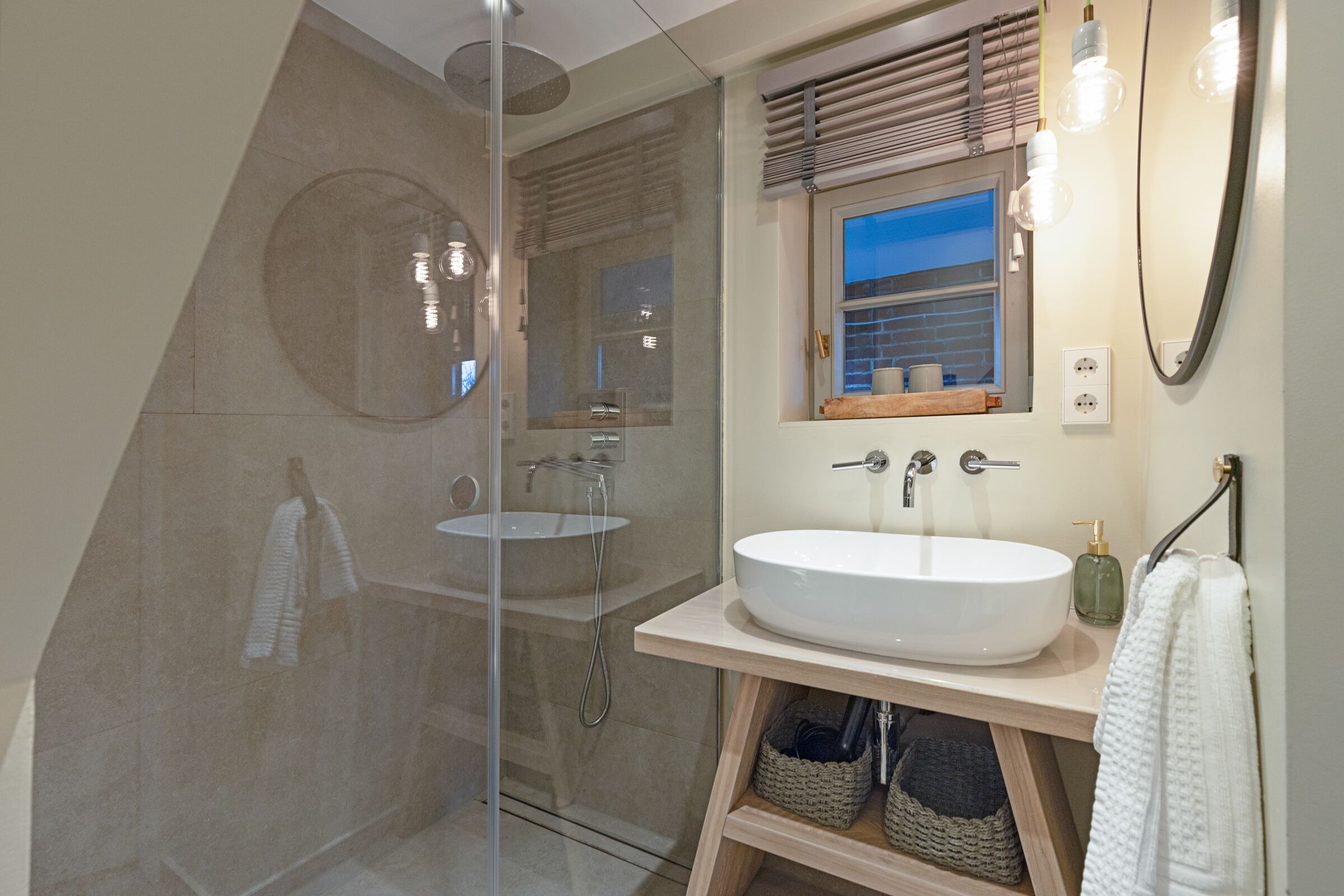 Un baño moderno con ducha de vidrio, dos lavamanos, tocador de madera y una ventana con persianas.