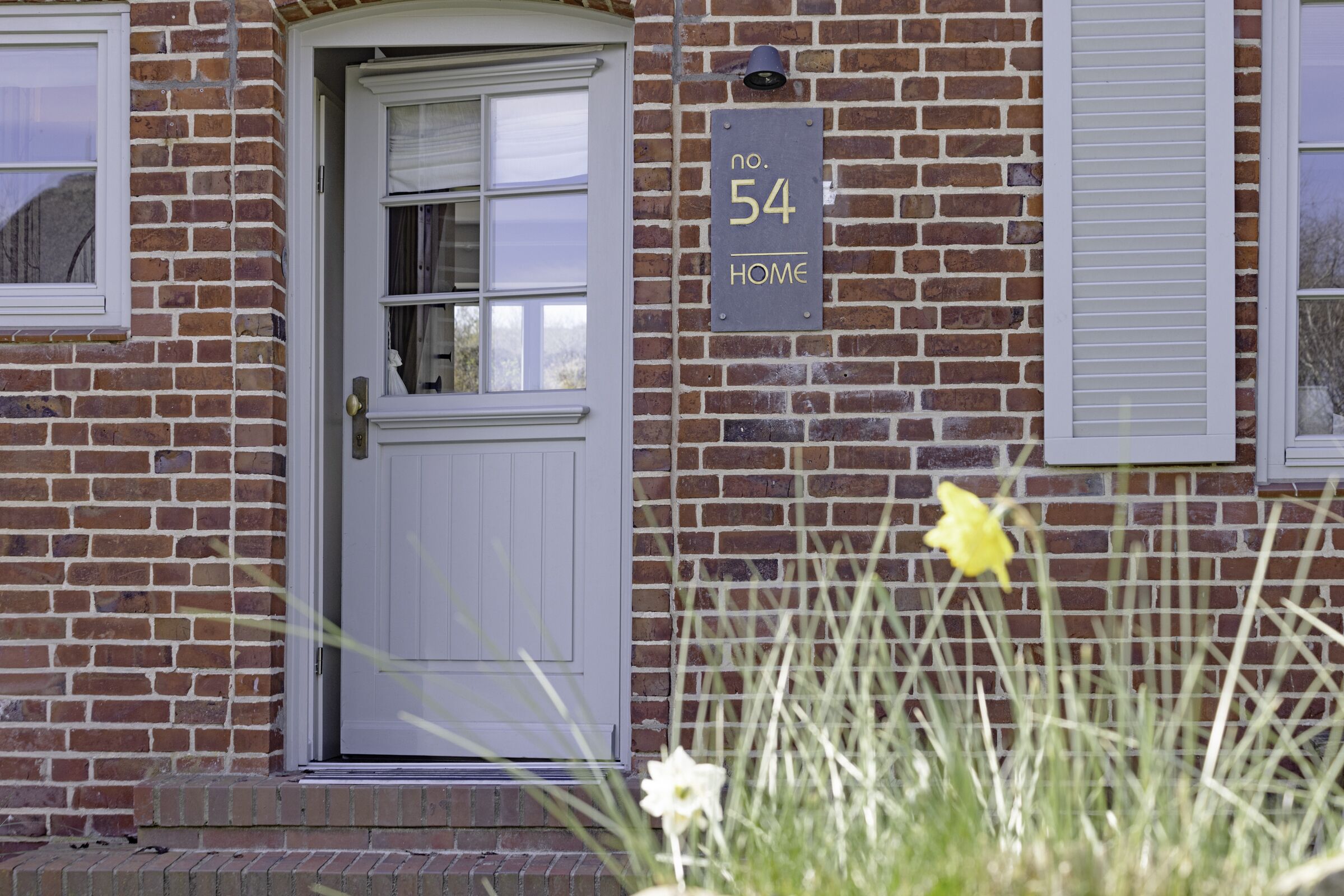 Een bakstenen huis met een witte deur en luiken, een bordje met 'nr. 54 HOME', en een narcis op de voorgrond.