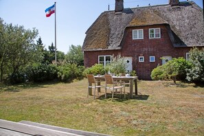 Ein Haus mit einem Strohdach und einer Ziegelaußenwand, eine Fahnenstange mit einer Flagge sowie ein Tisch mit Stühlen im Vordergrund.