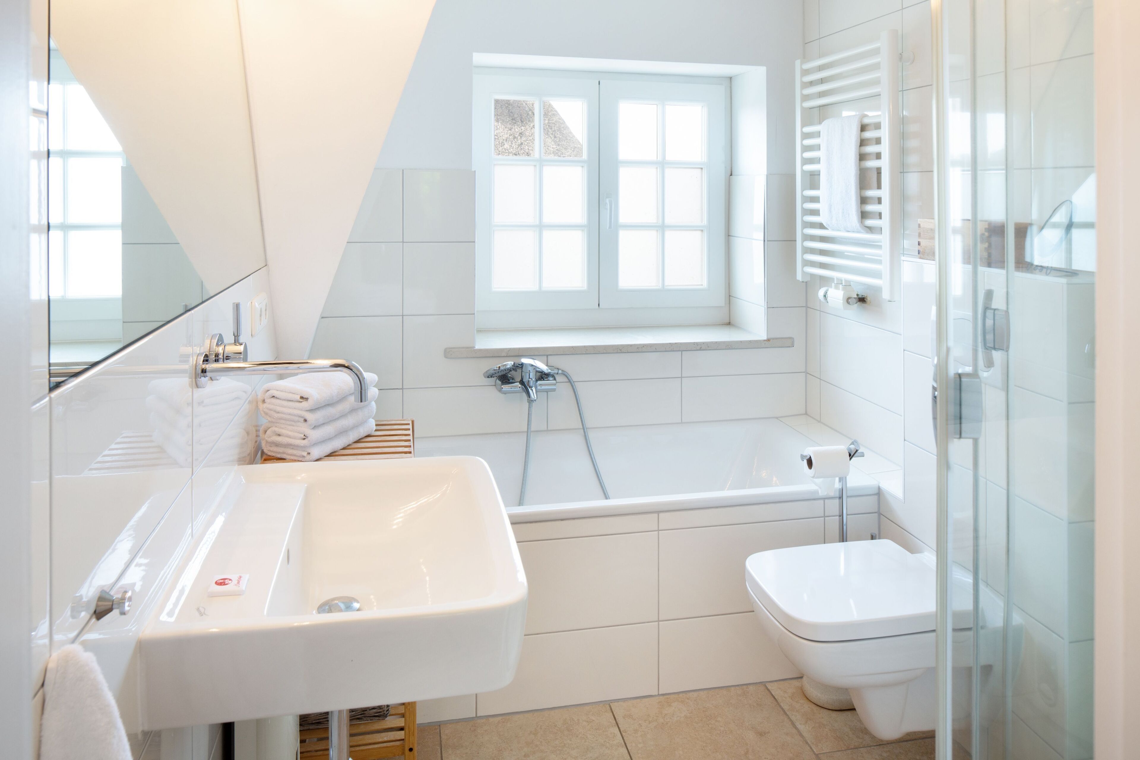 A modern bathroom with a white sink, toilet, and bathtub. There is a window above the bathtub, and a towel rack with towels is visible.