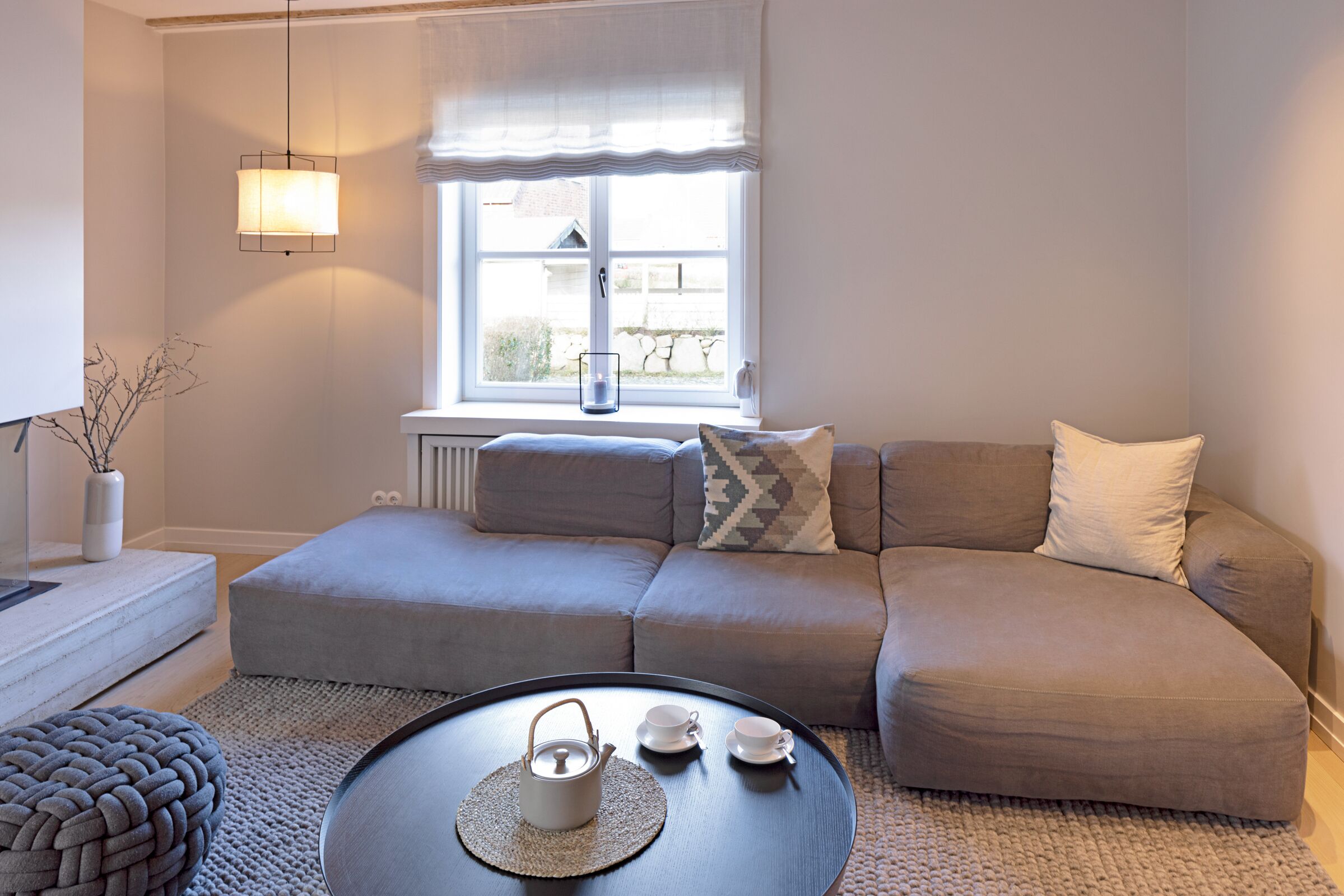 Una sala de estar moderna con un sofá gris, una mesa de centro redonda con una tetera, tazas y un tapete tejido. Se ve una ventana con vistas al exterior, una lámpara y un florero con ramas sobre una mesita auxiliar.