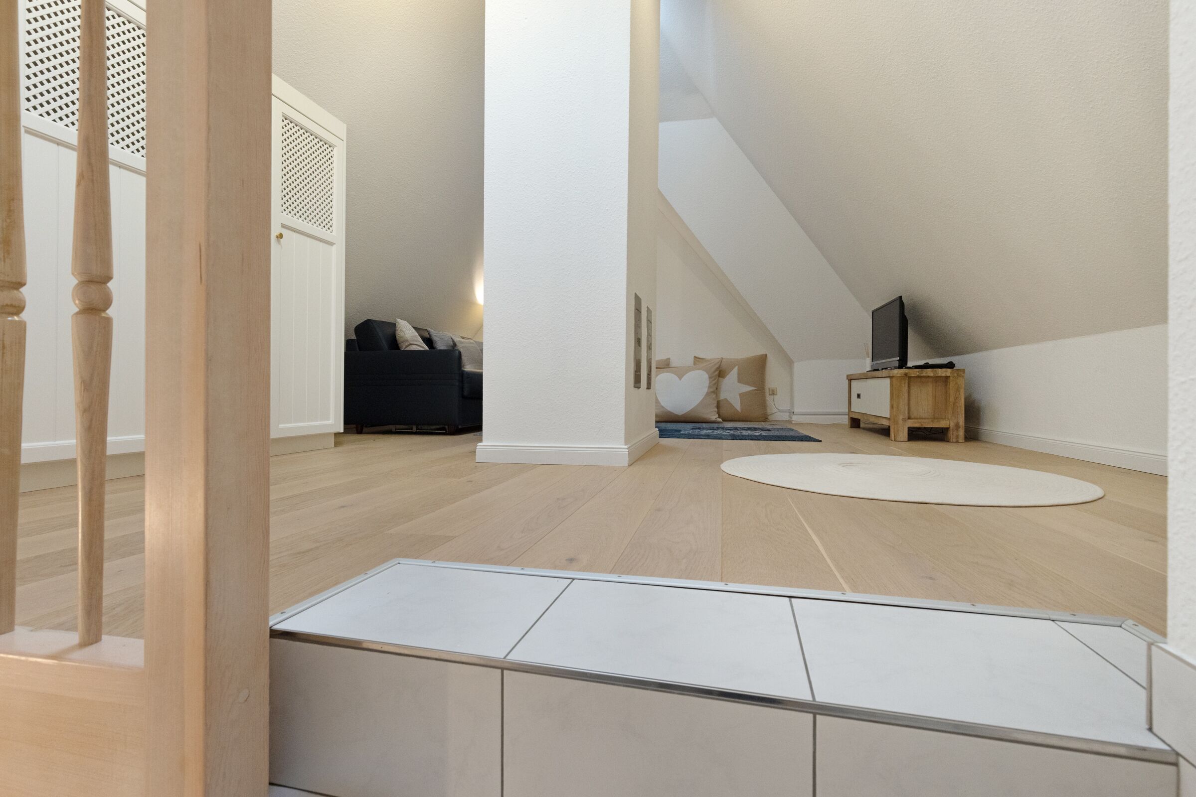 Een ruime slaapkamer op zolder met een schuine kap, een houten bed, een kleine televisie op een houten standaard en een witte vloerkleed.