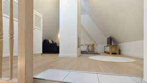 Un dormitorio amplio en el ático con techo inclinado, una cama de madera, un televisor pequeño sobre un soporte de madera y una alfombra blanca en el suelo.