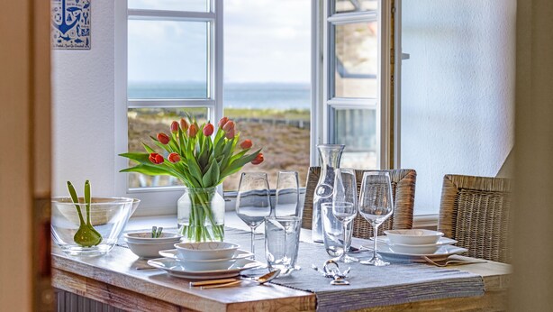 Una mesa de comedor dispuesta con copas y platos, un florero con tulipanes rojos, y vista al mar a través de amplias ventanas.