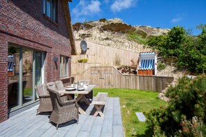 A patio with wicker furniture and a wooden table, a brick building, and a wooden fence. - Herzlich Willkommen im Luxus-ferienhaus Düünkieker "ankommen, Wohlfühlen!" (List)