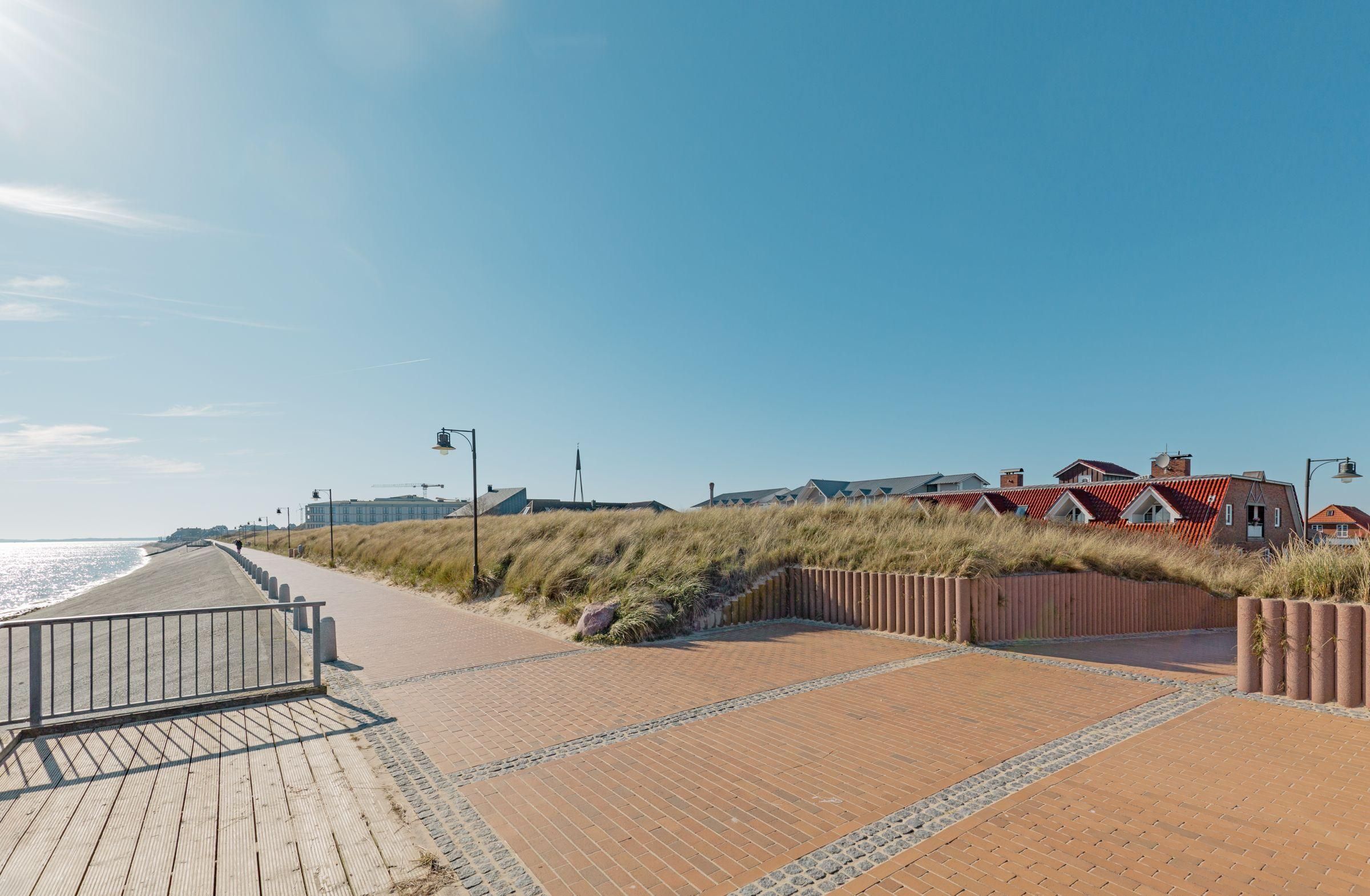 Une promenade en bois qui mène à une plage bordée de dunes, avec des bâtiments visibles à l’arrière-plan.