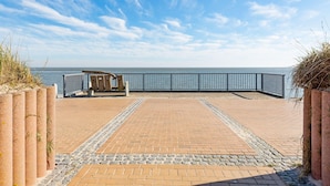 Ein gepflasterter Weg führt zu einem Strand, mit Metallgeländer und einer hölzernen Bank.