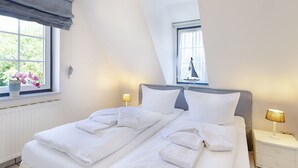 Ein ordentlich bezogenes Doppelbett mit weißen Bettwäsche und Handtüchern, ein Nachttisch mit Lampe, ein Fenster mit Blick auf Grünflächen und ein kleines dekoratives Segelboot.