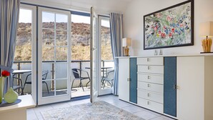 Ein Zimmer mit einer Schiebetür zu einer Terrasse, einem weißen Schrank mit Schubladen, einem floralen Gemälde und einem kleinen Tisch mit Vase und Kerze.