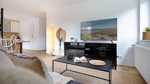 Ein modernes Wohnzimmer mit einem großen Flachbildfernseher, einem Essbereich mit Tisch und Stühlen sowie einer Küche im Hintergrund.