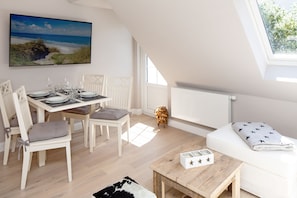 A dining area with a table set for four, a white sofa, a small wooden table, and a large window with a view of the beach.