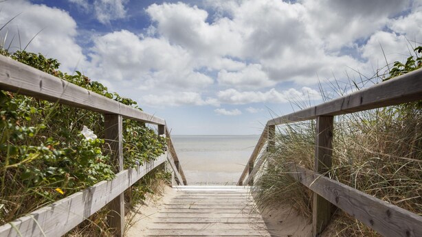 Ein hölzerner Steg führt zu einem Strand, der von einem klaren Himmel mit Wolken gesäumt wird.
