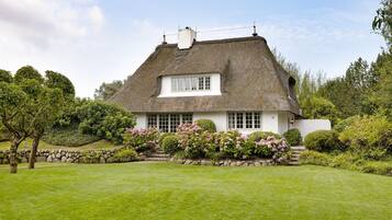 A thatched-roof house with a white facade, surrounded by a well-maintained garden with flowering plants and a lawn.