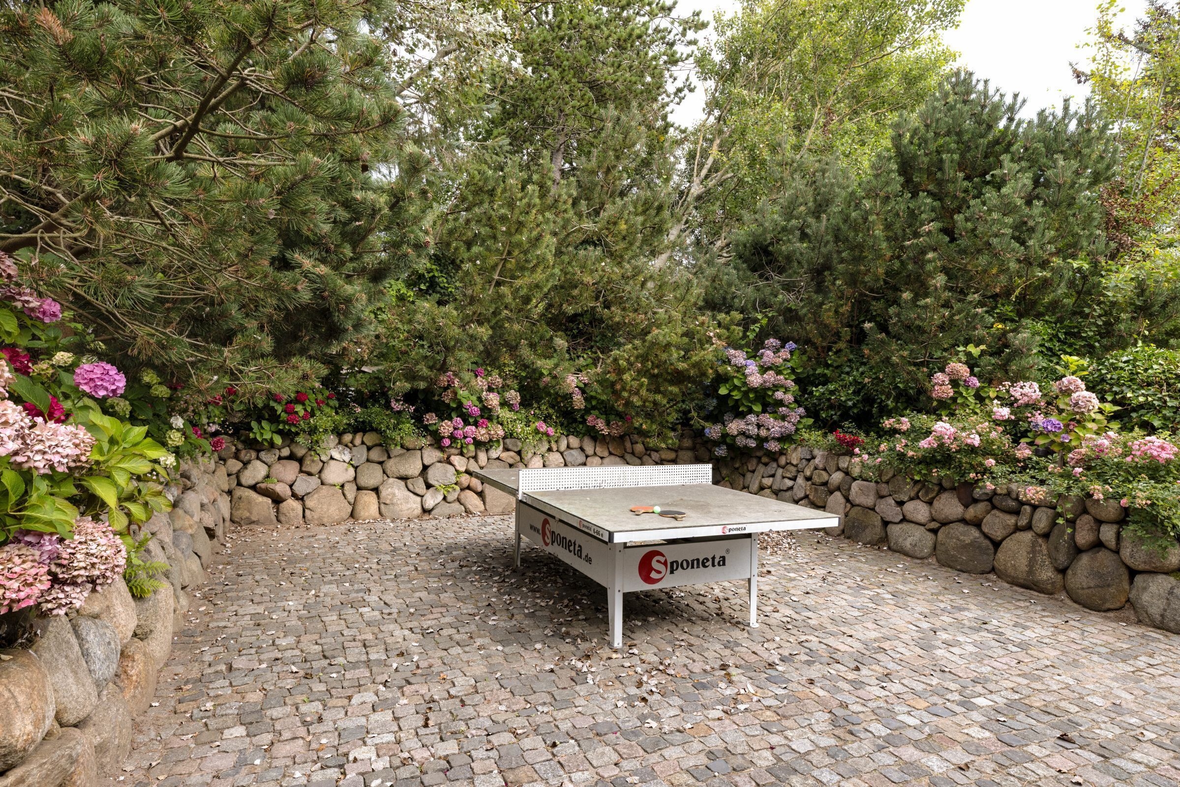 A ping pong table in an outdoor garden area with a stone wall and various plants.